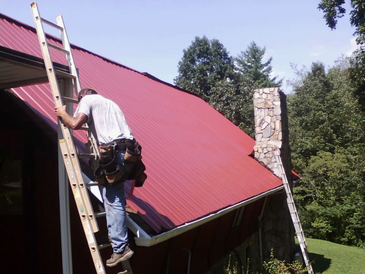 Roofer on a ladder beside a red metal roof and stone chimney — Soffit & Fascia Repair by Roofing Brothers in Porter Corners